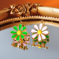 Golden Twin Daisy Hoop Earrings featuring one green enamel daisy and one soft pink enamel daisy suspended from polished gold-tone hoops.